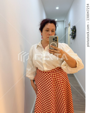 Woman taking mirror selfie in white short sleeve blouse and red gingham midi skirt at home. Outfit coordination, wardrobe styling, everyday elegance, personal fashion documentation, feminine clothing 138047254