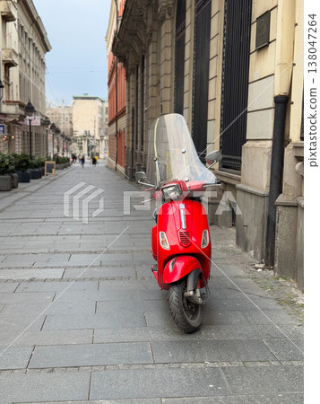 Red motor scooter standing alone on a narrow paved street between historic urban facades in daylight. Personal transportation, city navigation, independent travel, compact mobility, metropolitan 138047264