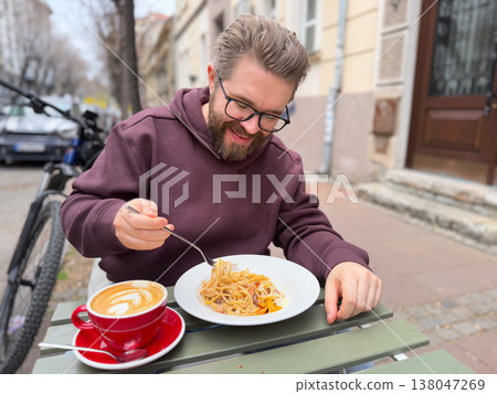 Bearded man twirling spaghetti with fork while sitting at an outdoor cafe table beside a red latte cup. Street dining ritual, relaxed meal break, urban cafe culture, approachable everyday lifestyle Bearded man twirling spaghetti with fork while sitting at an outdoor cafe table beside a red latte cup. Street dining ritual, relaxed meal break, urban cafe culture, approachable everyday lifestyle 138047269