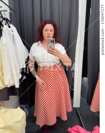 Woman taking mirror selfie in fitting room wearing white blouse with red gingham midi skirt. Outfit coordination, boutique styling, garment comparison, wardrobe planning, personal fashion selection 138047277