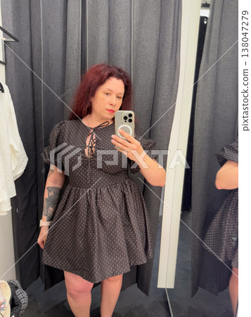 Woman taking mirror selfie in fitting room wearing black mini dress with dotted fabric. Dress fitting, fashion purchase, boutique browsing, outfit evaluation, personal style experimentation in retail 138047279