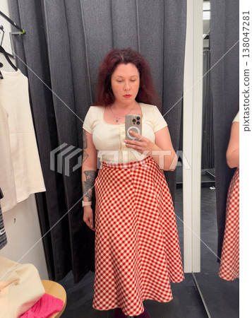 Woman taking mirror selfie in fitting room wearing cream knit top and red gingham midi skirt. Everyday fashion styling, garment fitting, retail decision making, personal wardrobe selection Woman taking mirror selfie in fitting room wearing cream knit top and red gingham midi skirt. Everyday fashion styling, garment fitting, retail decision making, personal wardrobe selection 138047281