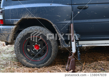 Hunting rifle leaning against a muddy off-road truck wheel after a rural field hunt 138047839