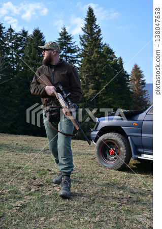Bearded hunter carrying a scoped rifle beside a 4x4 truck in a forest field Bearded hunter carrying a scoped rifle beside a 4x4 truck in a forest field 138047858