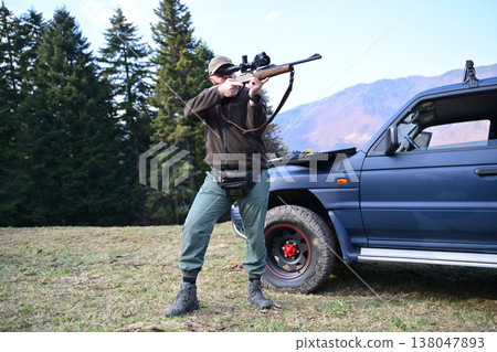 Hunter aiming rifle beside a parked SUV in mountainous forest landscape during a hunting trip Hunter aiming rifle beside a parked SUV in mountainous forest landscape during a hunting trip 138047893