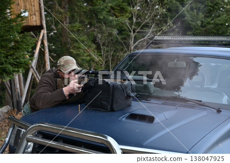 Hunter taking aim with a scoped rifle from a pickup truck hood in a forested setting Hunter taking aim with a scoped rifle from a pickup truck hood in a forested setting 138047925