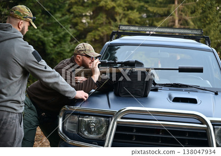 Two men in camouflage using a scoped rifle on a vehicle hood for tactical outdoor shooting practice 138047934