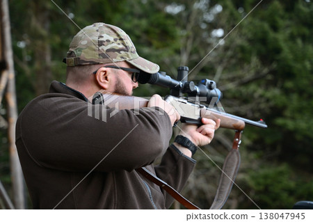 Hunter aiming a scoped rifle while wearing a camouflage cap in a forest setting Hunter aiming a scoped rifle while wearing a camouflage cap in a forest setting 138047945
