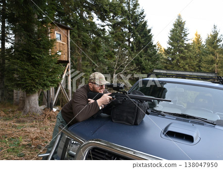 Hunter aiming scoped rifle from pickup truck hood near a wooden hunting blind in evergreen forest Hunter aiming scoped rifle from pickup truck hood near a wooden hunting blind in evergreen forest 138047950