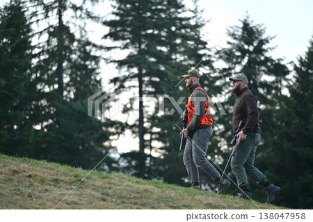 Two hunters walking through forest hillside carrying rifles and wearing hunting gear and orange vest 138047958
