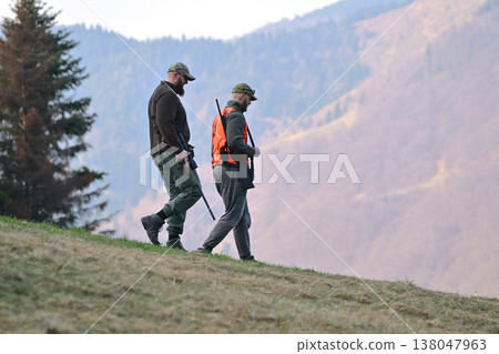 Two hunters walking down a mountain slope carrying rifles during autumn hunting trip Two hunters walking down a mountain slope carrying rifles during autumn hunting trip 138047963
