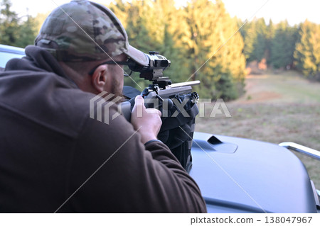 Man aiming a scoped rifle from truck bed for target shooting and hunting in woodland field Man aiming a scoped rifle from truck bed for target shooting and hunting in woodland field 138047967