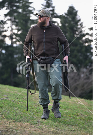 Bearded man carrying rifles walking on grassy hill in outdoor hunting gear and camouflage cap Bearded man carrying rifles walking on grassy hill in outdoor hunting gear and camouflage cap 138047973