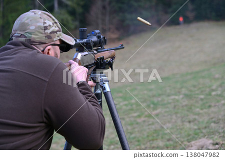Man shooting a scoped rifle on a tripod during outdoor target practice in a grassy field 138047982