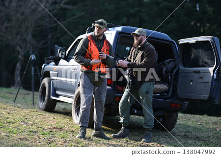 Two hunters preparing gear by an SUV at the edge of a forest during a daytime hunt Two hunters preparing gear by an SUV at the edge of a forest during a daytime hunt 138047992