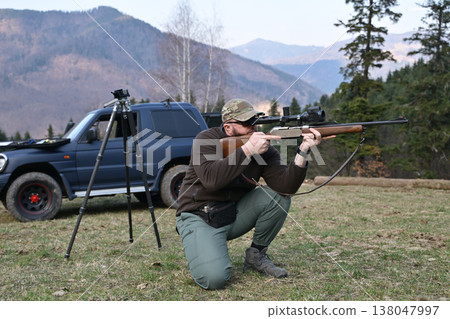 Hunter kneeling and aiming a scoped rifle in mountain field next to SUV and tripod Hunter kneeling and aiming a scoped rifle in mountain field next to SUV and tripod 138047997