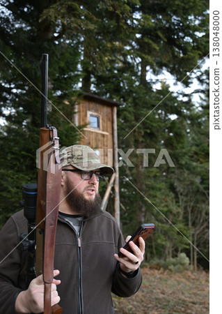 Hunter with rifle and smartphone near a wooden hunting blind in a forested outdoor setting Hunter with rifle and smartphone near a wooden hunting blind in a forested outdoor setting 138048000