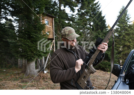 Hunter adjusting scoped rifle near a wooden deer stand in dense evergreen woods 138048004