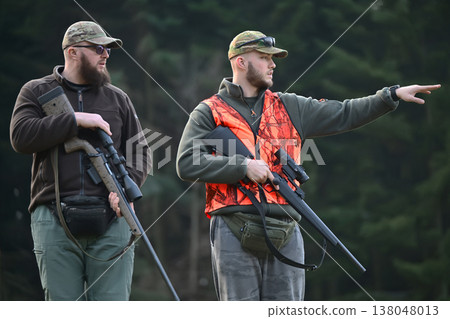 Two hunters in camo and blaze orange vest with rifles pointing and scouting in forest Two hunters in camo and blaze orange vest with rifles pointing and scouting in forest 138048013