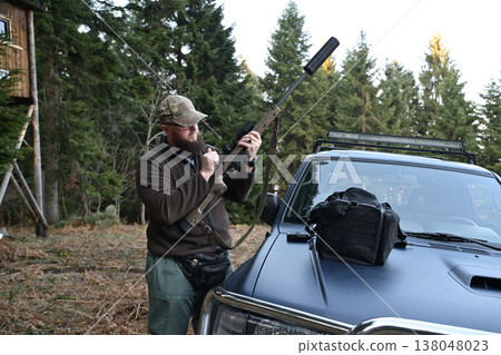 Hunter preparing a scoped rifle beside an off-road vehicle in a forested hunting camp 138048023