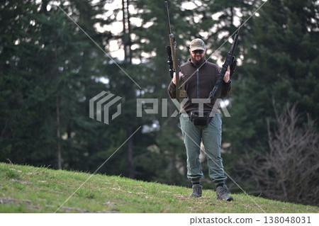 Bearded hunter carrying two rifles walks on grassy hillside in forest during hunting trip Bearded hunter carrying two rifles walks on grassy hillside in forest during hunting trip 138048031