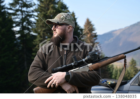 Bearded hunter with rifle and scope stands in forest clearing wearing camouflage cap and outdoor gear 138048035