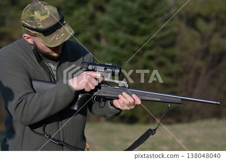 Hunter adjusting scope on a bolt-action rifle at a woodland shooting range during daytime Hunter adjusting scope on a bolt-action rifle at a woodland shooting range during daytime 138048040
