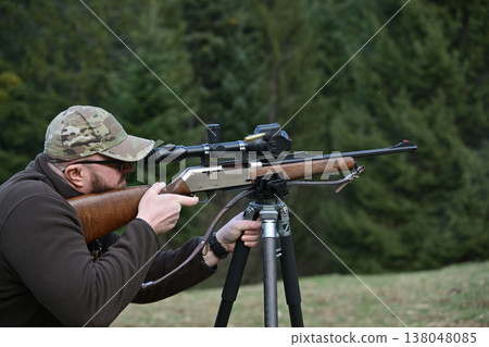 Hunter aiming a scoped rifle from a tripod in a forest clearing during outdoor hunting practice 138048085