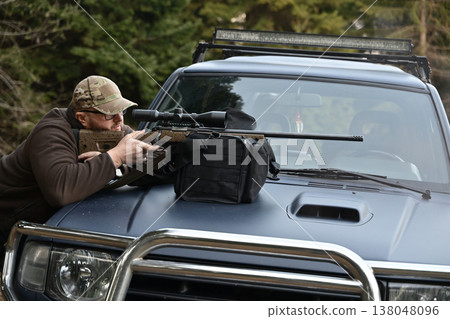 Man aiming scoped rifle from hood of pickup truck in forested outdoor shooting position Man aiming scoped rifle from hood of pickup truck in forested outdoor shooting position 138048096