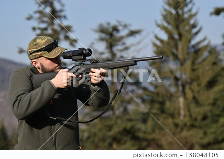Hunter aiming rifle with scope in forest clearing wearing camouflage cap and outdoor clothing Hunter aiming rifle with scope in forest clearing wearing camouflage cap and outdoor clothing 138048105