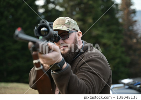 Bearded hunter aiming a scoped rifle in the woods wearing camouflage cap and outdoor jacket 138048133