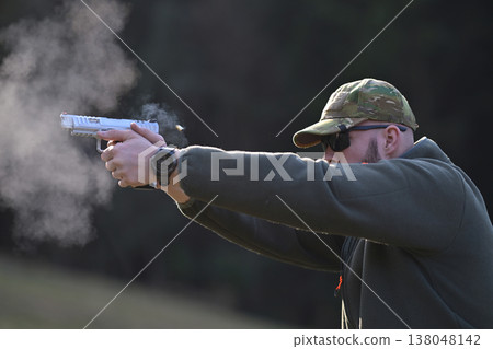 Man firing handgun at outdoor range wearing camouflage cap and sunglasses with visible muzzle smoke 138048142
