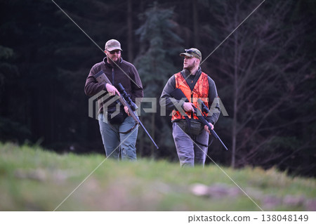 Two hunters walking through a forest field carrying rifles with one wearing a blaze orange vest Two hunters walking through a forest field carrying rifles with one wearing a blaze orange vest 138048149