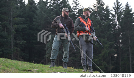 Two hunters standing with rifles in a forest wearing orange safety vest and camouflage caps 138048159