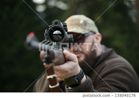 Hunter aiming a scoped rifle downrange during outdoor target practice in camouflage cap and sunglasses 138048176