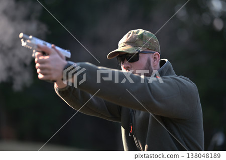 Man practicing handgun shooting outdoors in tactical gear and camouflage cap during target training session 138048189
