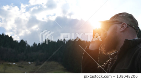 Bearded man in camo hat looking through binoculars at sunset over forested hills 138048206
