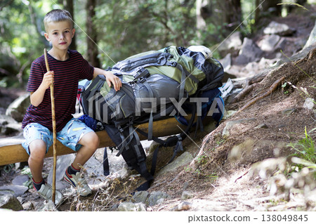 Young cute child boy with stick sitting alone at big tourist backpack on lit by bright summer sun rough wooden bench on green pine trees bokeh background. Tourism and active lifestyle concept. 138049845