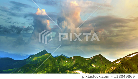 Wide panorama of lit by morning sun green valley, hills covered with forest and distant misty mountains with patches of snow under cloudy windy sky. Beauty of nature, tourism and traveling concept. Wide panorama of lit by morning sun green valley, hills covered with forest and distant misty mountains with patches of snow under cloudy windy sky. Beauty of nature, tourism and traveling concept. 138049869