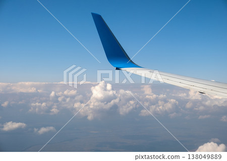 View of jet airplane wing from inside flying over white puffy clouds in blue sky. Travel and air transportation concept. 138049889