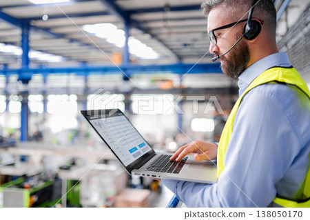 Side view of factory manager with headset and laptop standing in industrial factory. Side view of factory manager with headset and laptop standing in industrial factory. 138050170