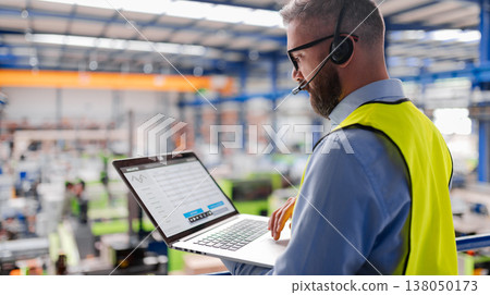 Side view of factory manager with headset and laptop standing in industrial factory. Side view of factory manager with headset and laptop standing in industrial factory. 138050173