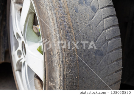 A car wheel shows a damaged tire with visible metal corner while parked on the roadside during the day. Signs of wear are present A car wheel shows a damaged tire with visible metal corner while parked on the roadside during the day. Signs of wear are present 138050850