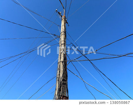 A utility pole stands in a city with many wires extending in different directions. The sky is blue with no clouds 138050867