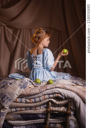 Little girl in blue dress with crown holding apple on mattresses, princess and the pea Little girl in blue dress with crown holding apple on mattresses, princess and the pea 138050895