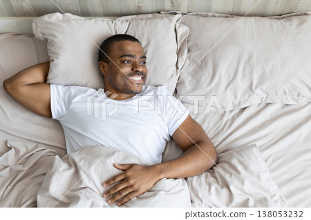 Black man lying in bed smiling and relaxed, looking away, representing comfort 138053232