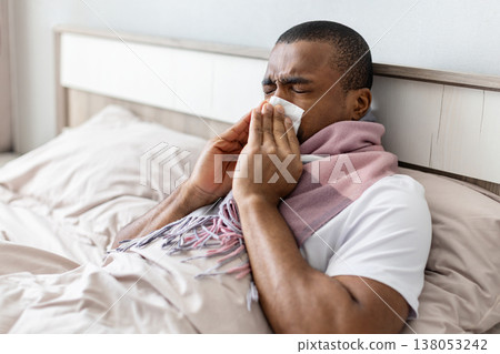 Young black man sitting in bed sneezing into tissue while wearing scarf Young black man sitting in bed sneezing into tissue while wearing scarf 138053242