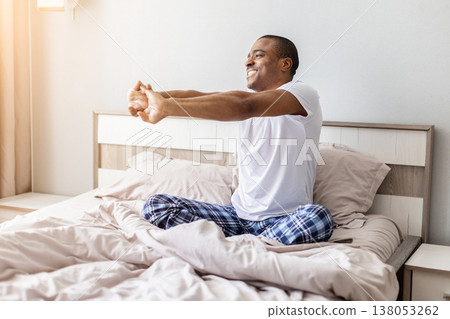 Young black man sitting in bed stretching arms forward after waking up 138053262