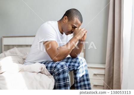 Upset black man sitting on bed with hands clasped and eyes closed 138053313