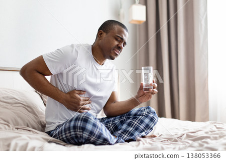 Young black man sitting on bed holding stomach and glass of water Young black man sitting on bed holding stomach and glass of water 138053366
